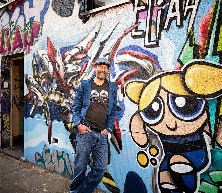 Künstler Adrian Savulescu vor einer Wand voller Graffitis in Köln-Kalk